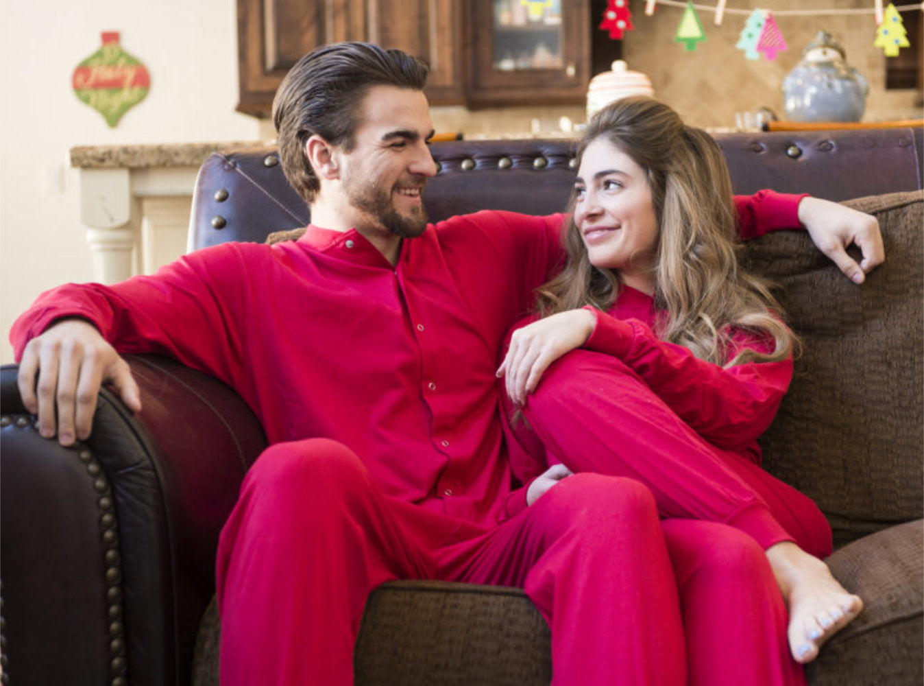 A couple sitting on the couch wearing matching red Cotton Knit Adult Footless Onesie Pajamas for Valentine's Day, from Big Feet PJs.