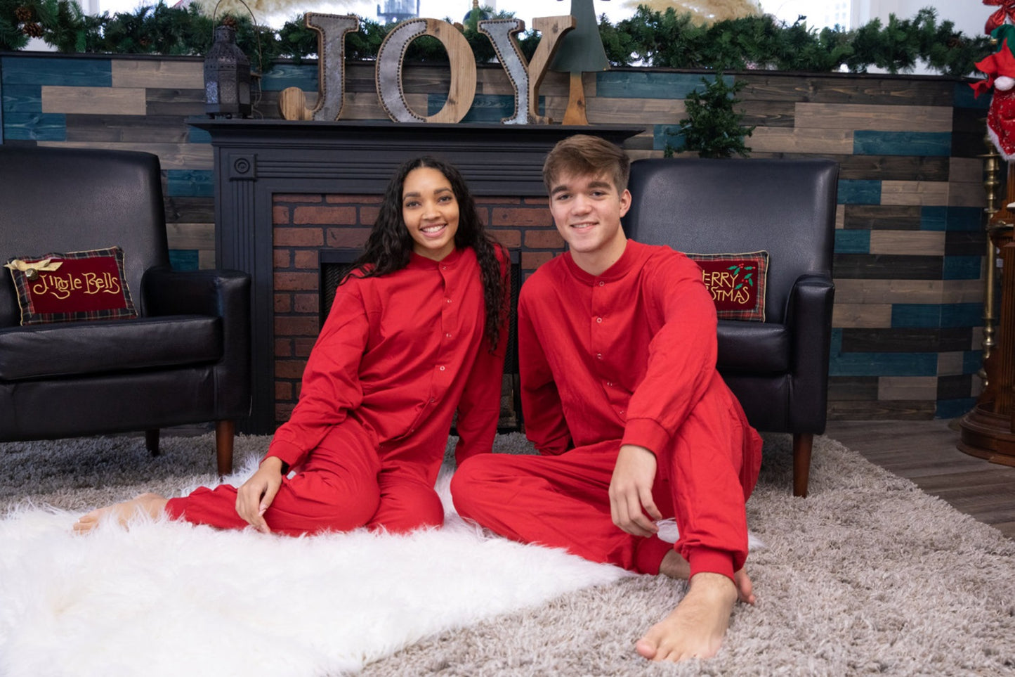 A couple by the fireplace for Christmas pictures, wearing matching unisex Red Cotton Knit Adult Footless Onesie Union-Suit style Pajamas from Big Feet Pajama Company.