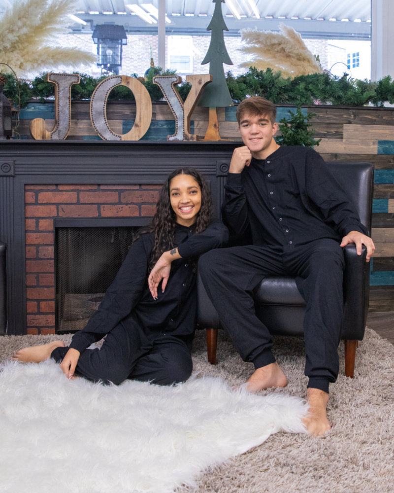 A couple by the fireplace for Christmas pictures, wearing matching unisex Black Cotton Knit Adult Footless Onesie Pajamas from Big Feet Pajama Company.