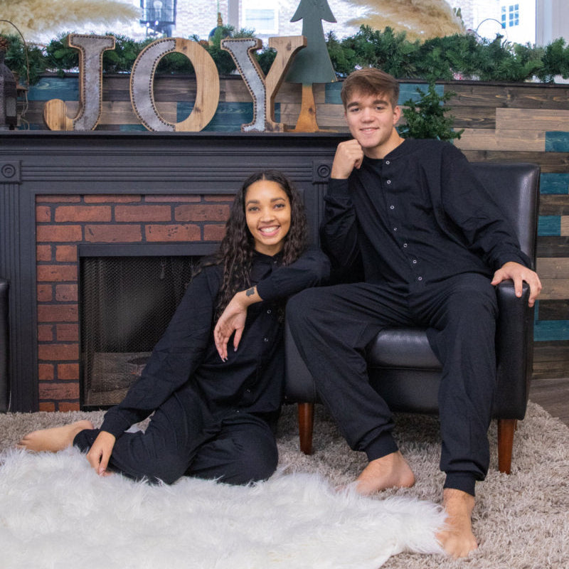 A couple by the fireplace for Christmas pictures, wearing matching unisex Black Cotton Knit Adult Footless Onesie Pajamas from Big Feet Pajama Company.