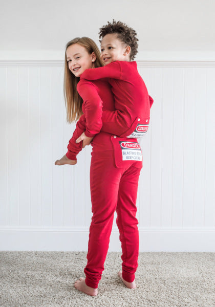 A girl is giving her little brother a piggyback ride. They are wearing matching red cotton union suit onesie pajamas adorned with a humorous butt flap that has a warning sign printed on them which reads "DANGER - BLASTING AREA - KEEP CLEAR" , from the cotton onesie collection at Big Feet Pajama Company.