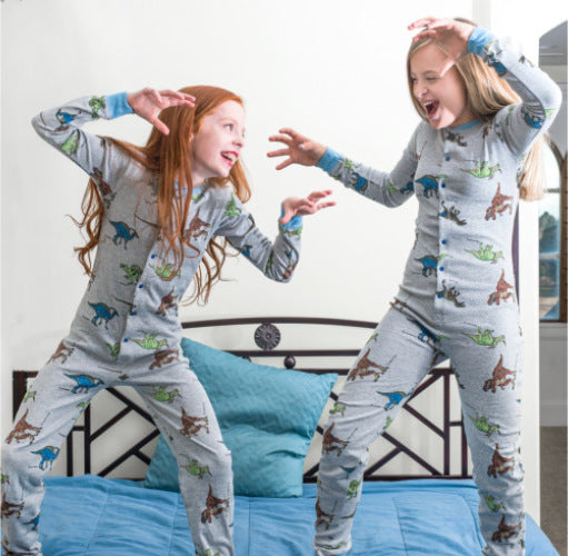 Two young girls are standing on their bed pretending to be T-Rex dinosaurs. They're wearing matching cotton footless onesies from Big Feet Pajama Company with five different realistic dinosaurs printed all over them.