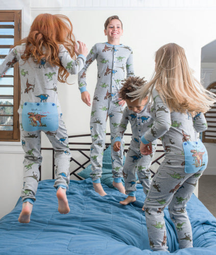 Four children celebrating Christmas Eve are all jumping on a bed and wearing matching dinosaur pajamas for Christmas. These matching pjs are heather blue cotton Union suit onesie pajamas adorned with 5 different dinosaurs printed all over them, from the cotton onesie collection at Big Feet Pajama Company.